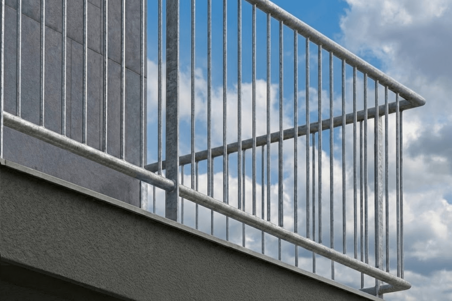 Modern verzinkt balkonhekwerk met een strak design van verticale spijlen op gelijke afstand, gemonteerd op een betonnen balkon tegen een grijze gevel.