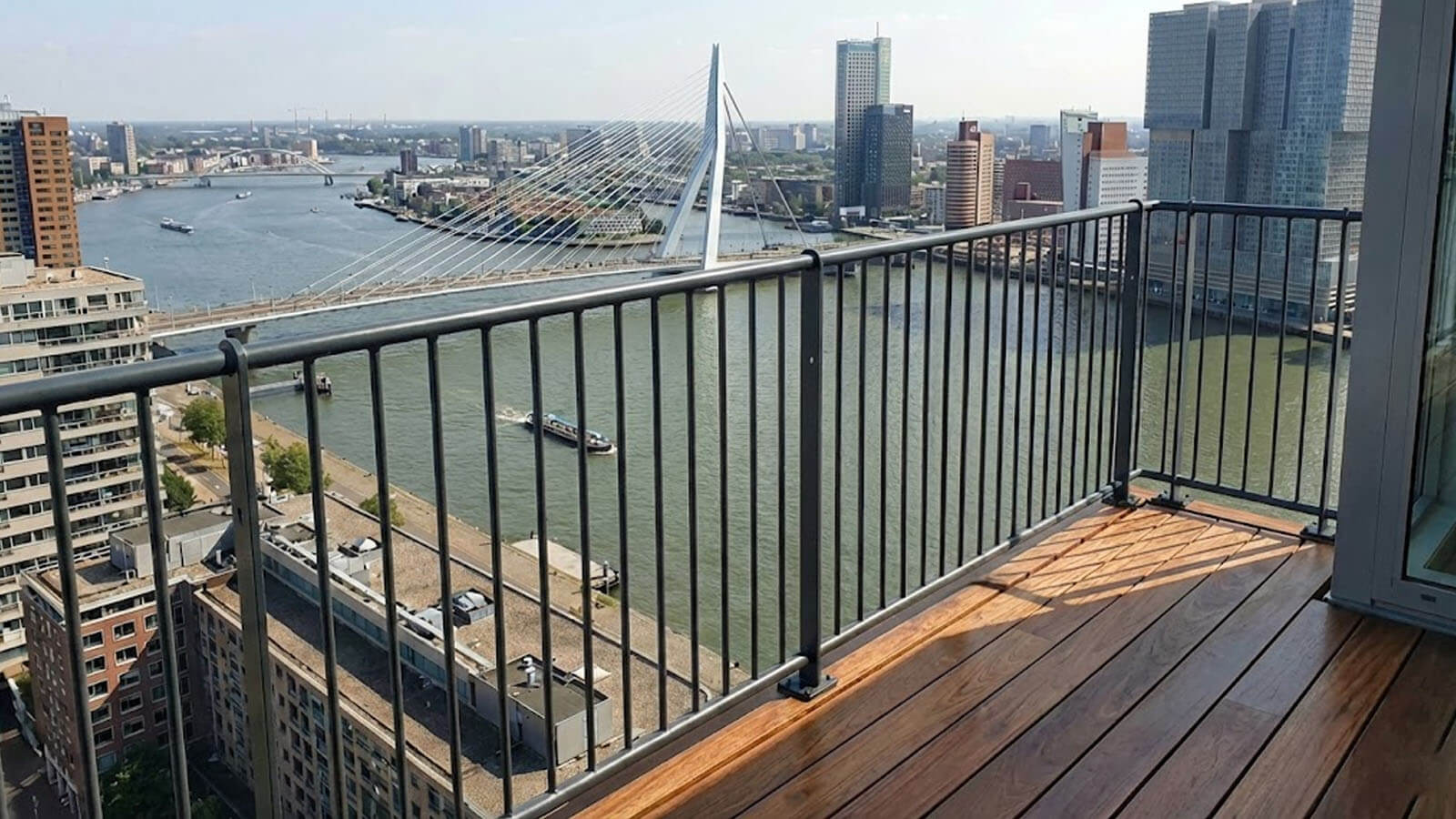 Modern antraciet spijlenhekwerk op een balkon met houten vlonders en uitzicht over de Erasmusbrug in Rotterdam.