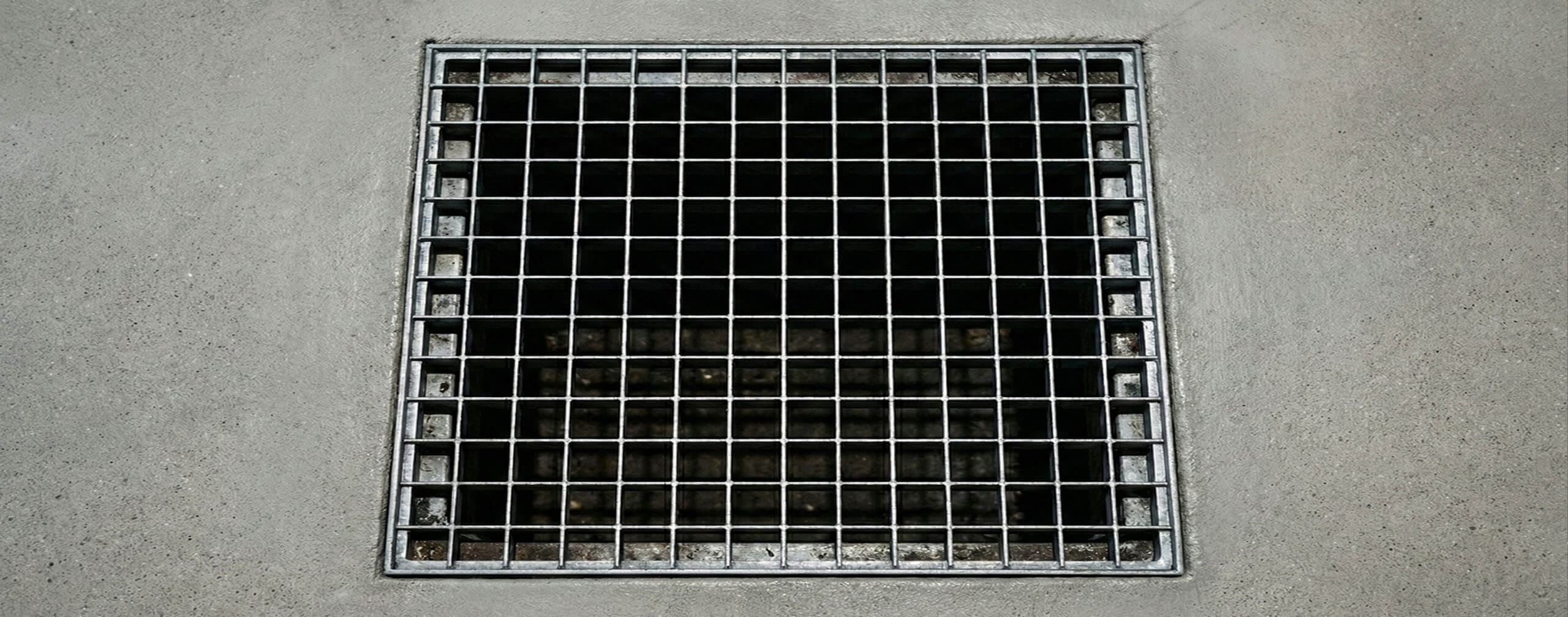 Bovenaanzicht van een verzinkt stalen persrooster met omlijsting, strak gemonteerd in een nieuw gestorte, lichtgrijze betonvloer.